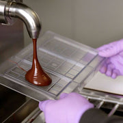 Brown chocolate dispensing out of silver pipe on to clear plastic chocolate block mold.