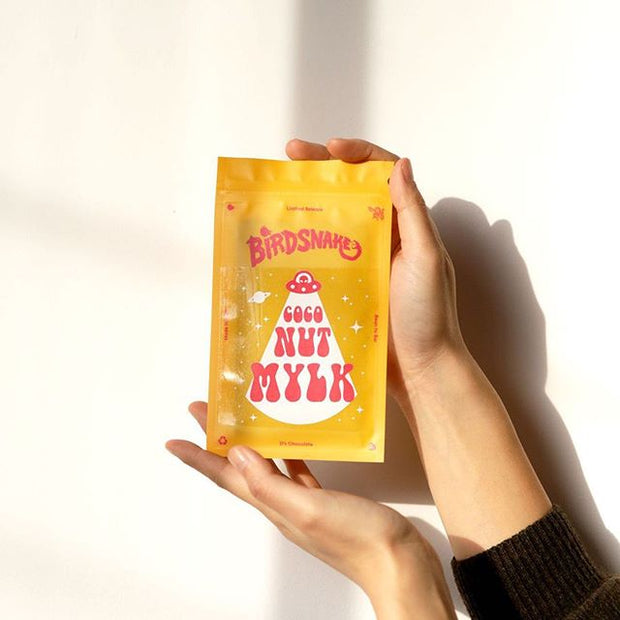 Woman holding a small yellow bag of birdnsake chocolate in front of white wall background.