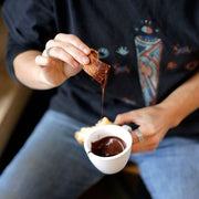 Woman dunking pastry into small white coffee mug filled with melted chocolate.
