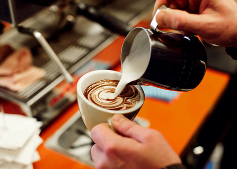 Barista pouring frothed milk from a silver milk jug into a hot chocolate.